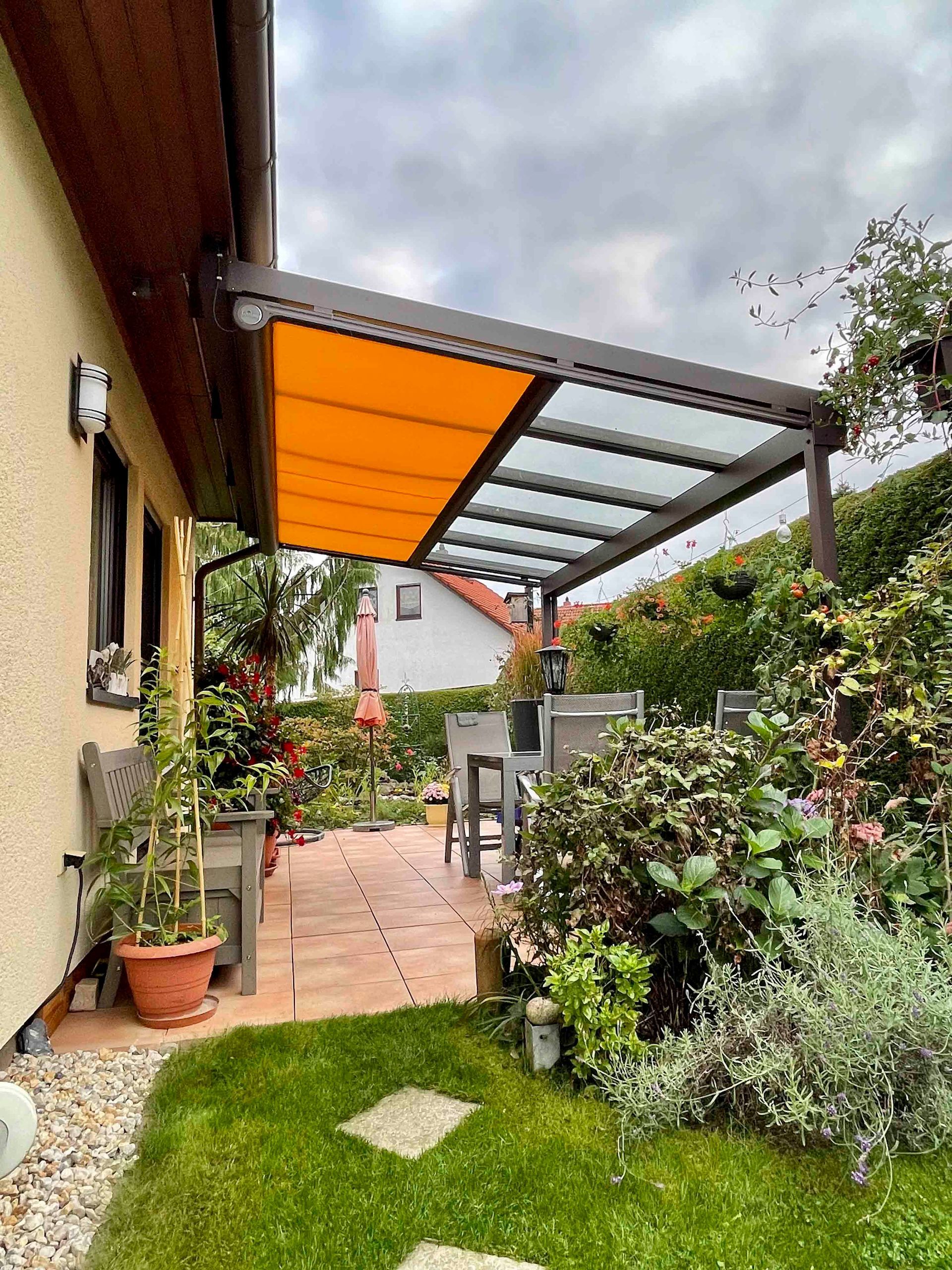 Terrasse mit orangefarbener und durchsichtiger Markise, Tisch, Stühlen, Pflanzen und Rasen.