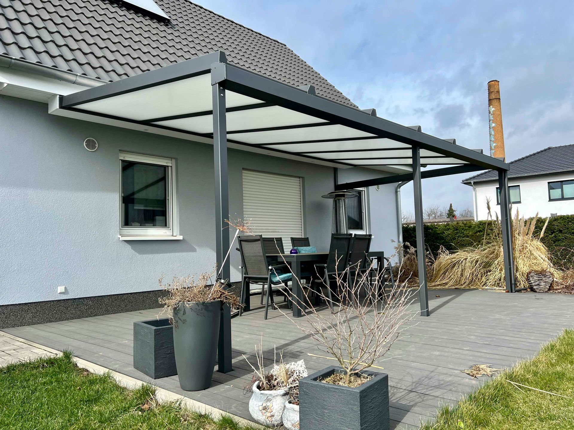 Graue Terrasse mit dunkelgrauer Pergola, Tisch und Stühlen, neben einem Haus.