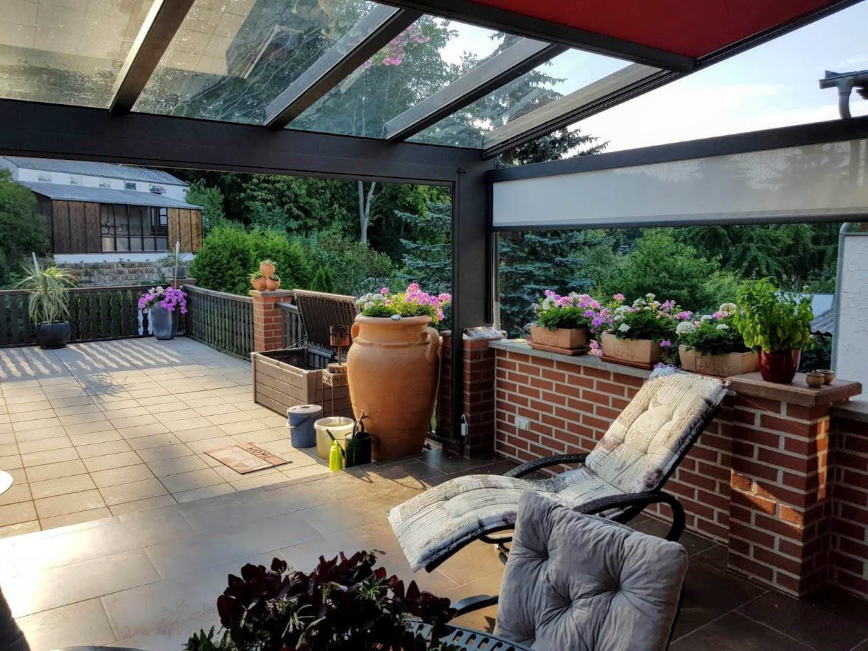 Terrasse mit Glasdach, Ziegelmauer und Liegestühlen, geschmückt mit Pflanzen und einer Urne; mit Blick auf den Garten.