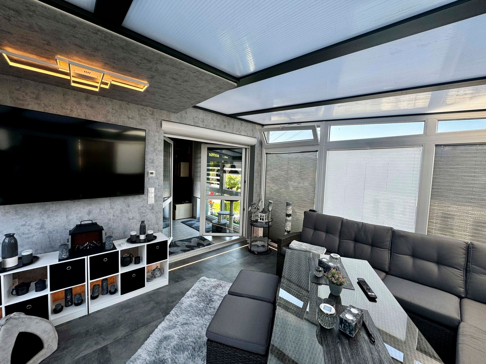 Moderner Wintergarten mit grauem Sofa, großem Fernseher und Glastisch, in der Nähe einer Tür nach draußen.