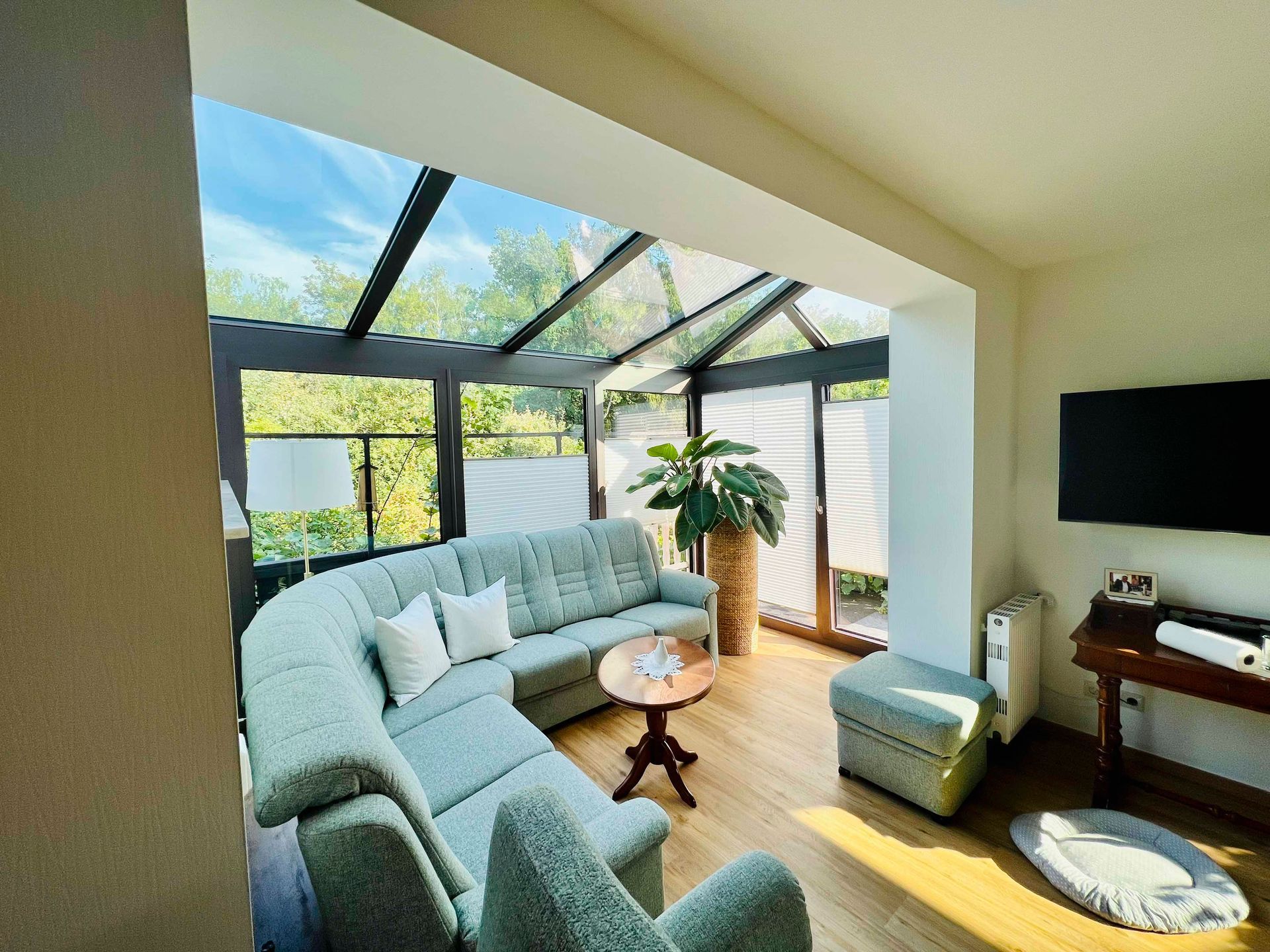 Wintergarten mit blaugrünem Sofa, Glasdach und Fernseher.