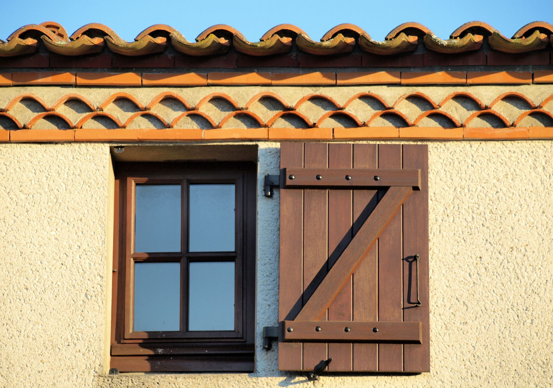Petite fenêtre et volet en bois brun sur un mur en crépi couleur crème, sous des tuiles en terre cuite