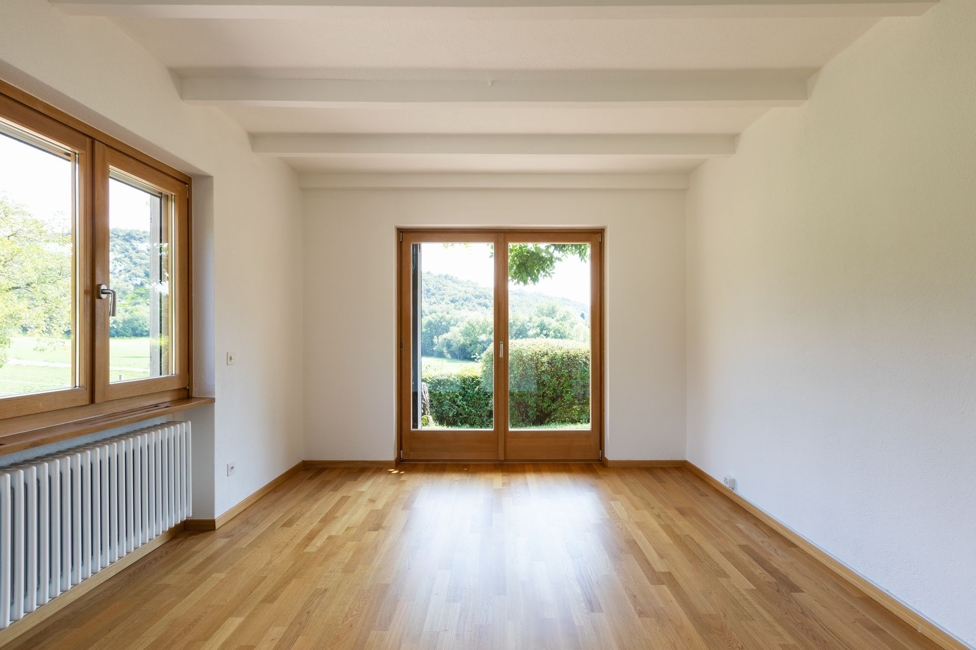 Pièce lumineuse et vide, avec un parquet, des murs blancs et de grandes fenêtres donnant sur la verdure