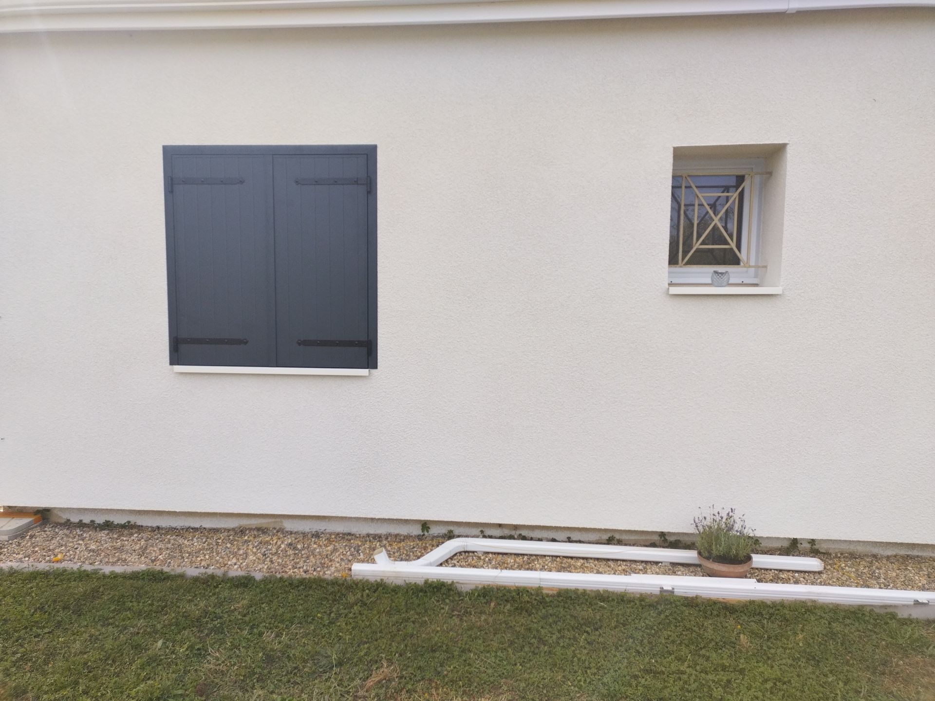 Mur extérieur blanc avec une fenêtre grise à volets, une petite fenêtre à barreaux et de l’herbe devant
