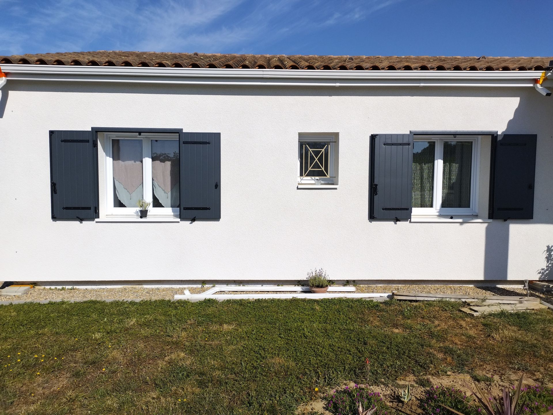 Extérieur d’une maison blanche avec deux fenêtres, des volets sombres et une petite bouche d’aération centrale donnant sur une pelouse