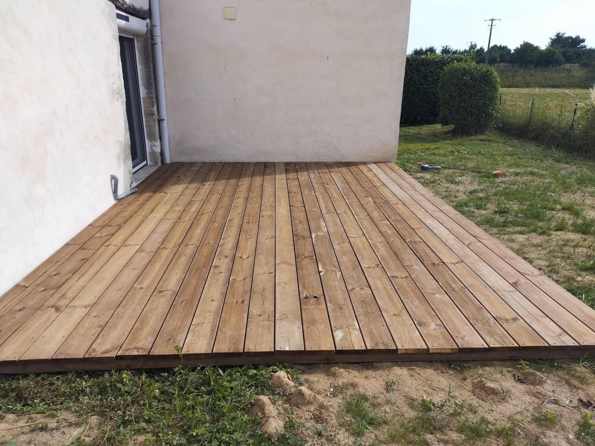 Terrasse en bois à côté d’une maison blanche, avec de l’herbe et un champ dégagé en arrière-plan.