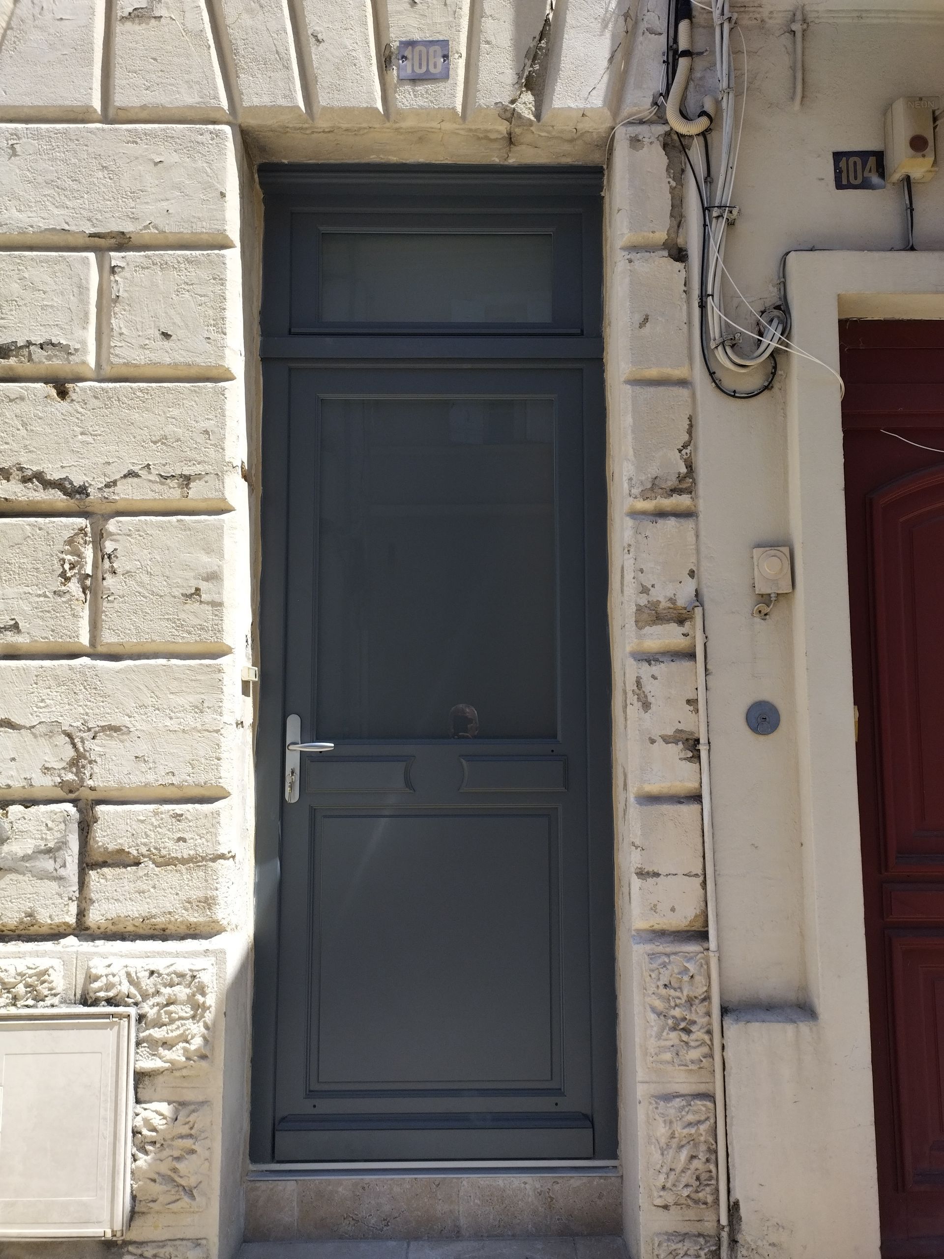 Porte d’entrée gris foncé intégrée dans un mur en pierre claire, à côté d’une porte rouge