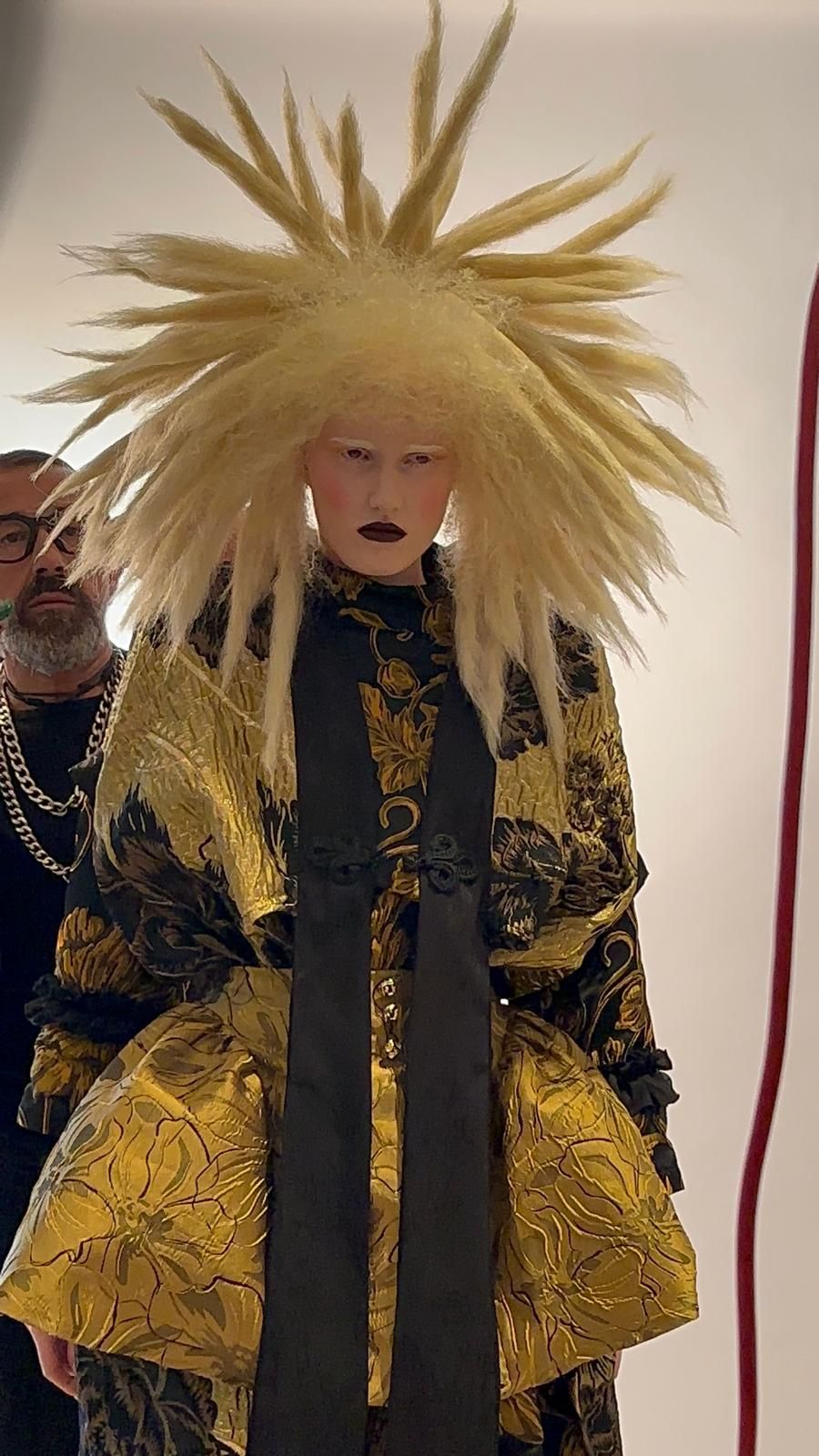 Persona con un elaborado vestido negro con estampado dorado y peinado rubio de punta. El hombre se ajusta el vestido.