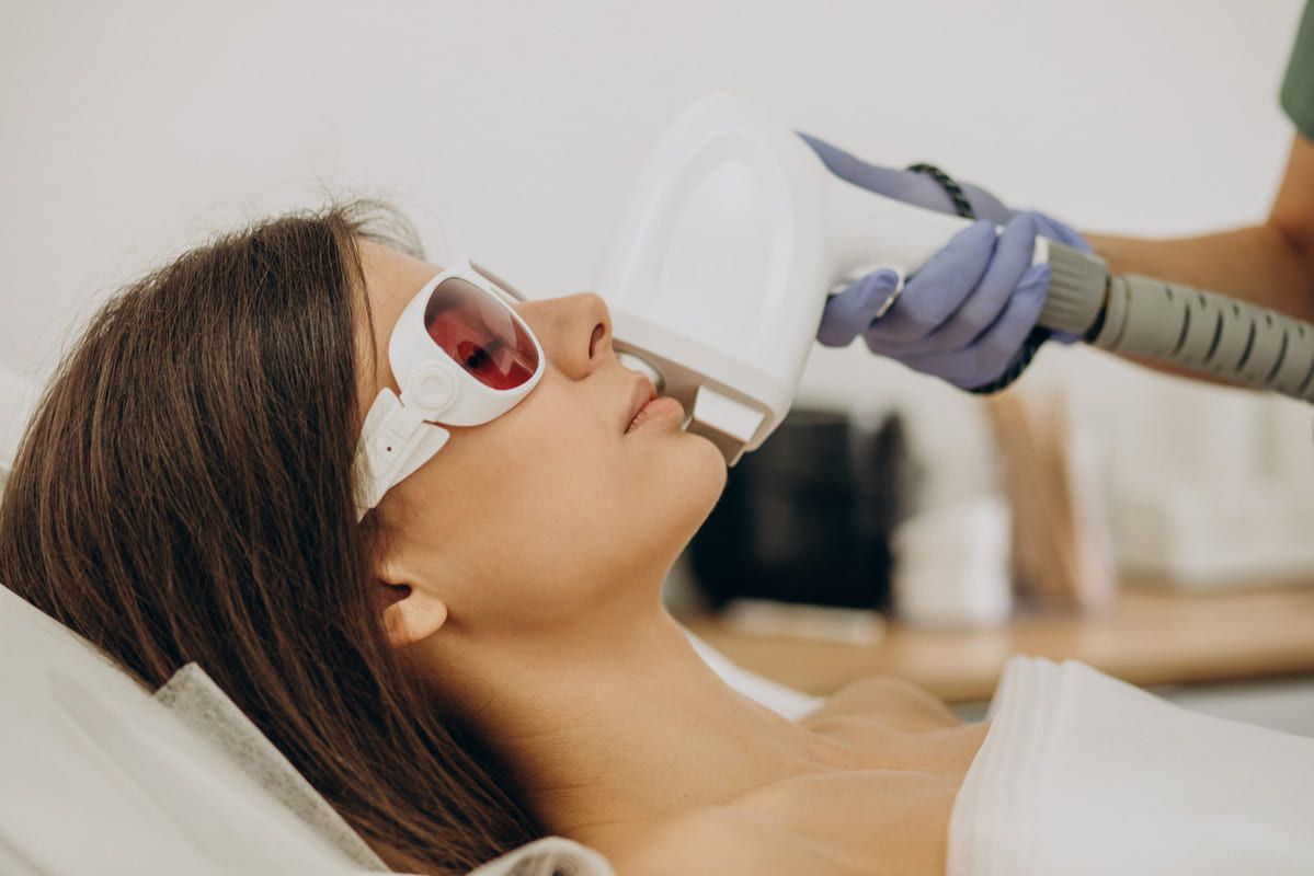 Paciente recibiendo un tratamiento facial con láser mientras está tumbada en una camilla de clínica y lleva gafas de protección