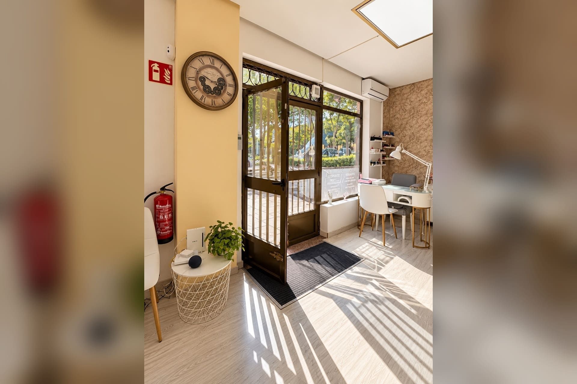 Entrada soleada de cafetería con puerta de cristal, reloj de pared, extintor rojo y una mesita junto a la ventana.