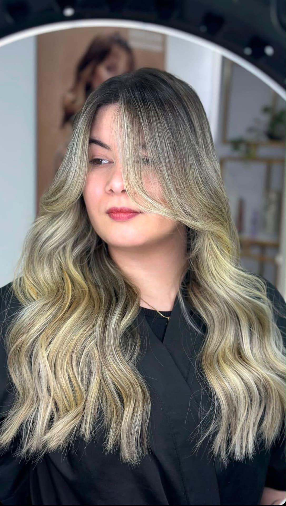 Mujer con el pelo largo, rubio y ondulado, con mechas plateadas, posando frente al espejo de un salón de belleza.