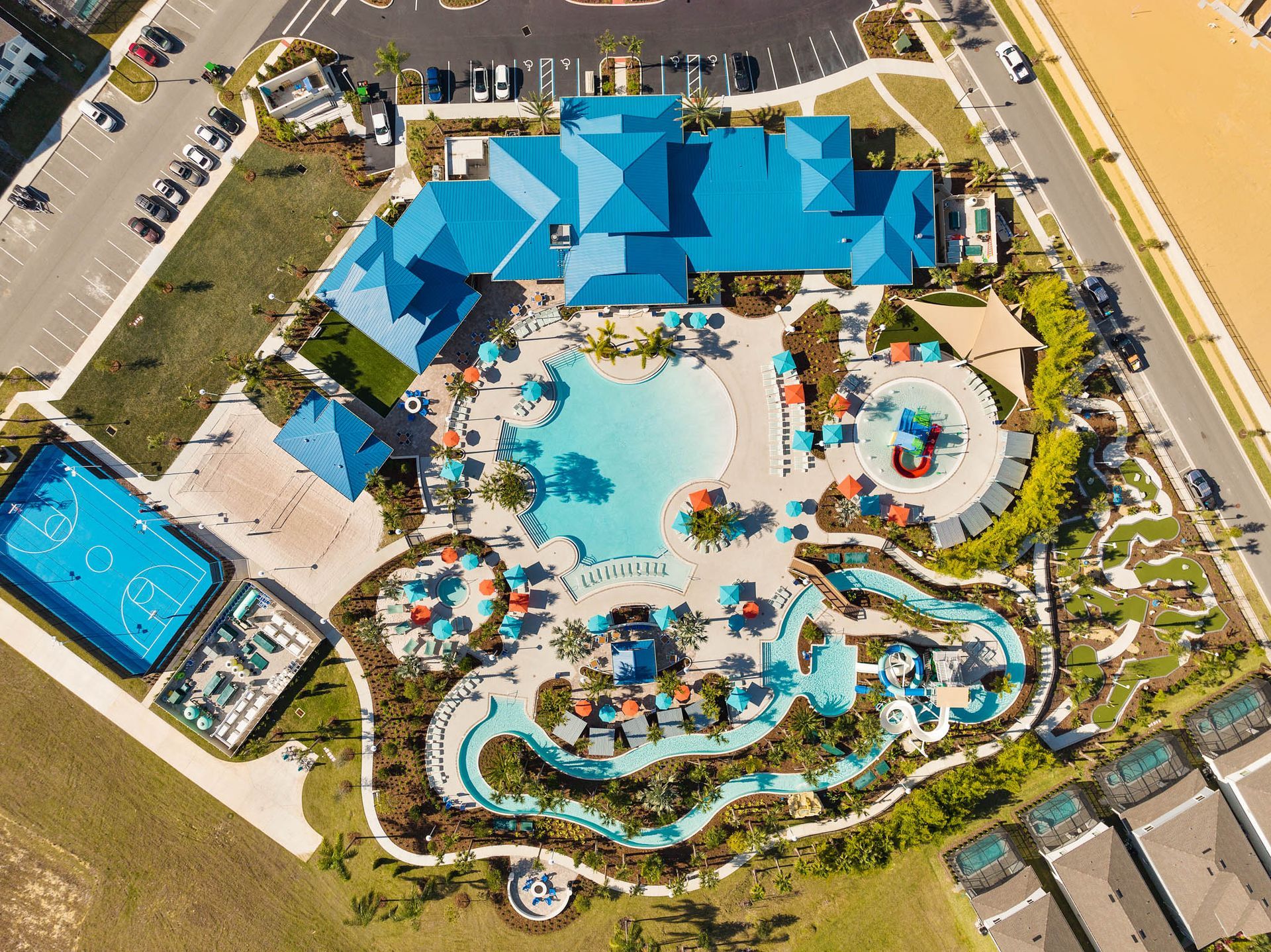 Aerial view: blue-roofed resort with swimming pools, basketball court, and buildings in a sunny setting.