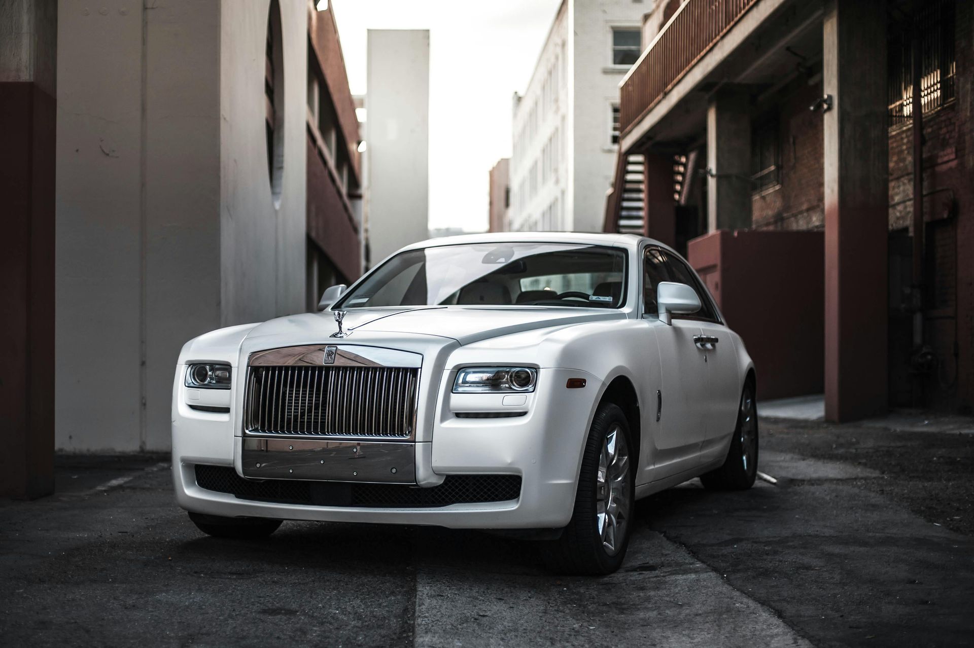 White Rolls-Royce sedan parked in a narrow alley between brick buildings, bright sunlight, front view.