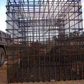 Gran jaula o caja de metal oxidado en una obra de construcción, con armazón de barras de refuerzo y un vehículo parcialmente visible.