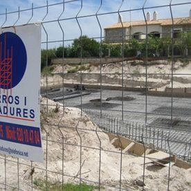 Obra en construcción detrás de una valla de alambre con zanjas de cimentación de hormigón y un letrero en el lado izquierdo.