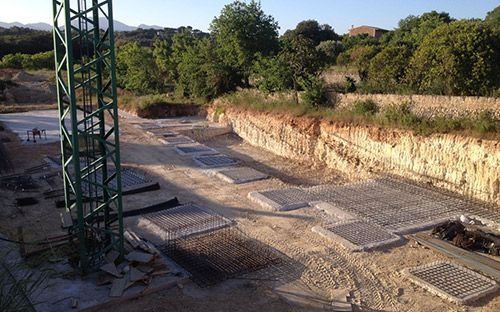 Obra en construcción con cimientos de hormigón, torre metálica y terraplén de tierra junto a árboles.