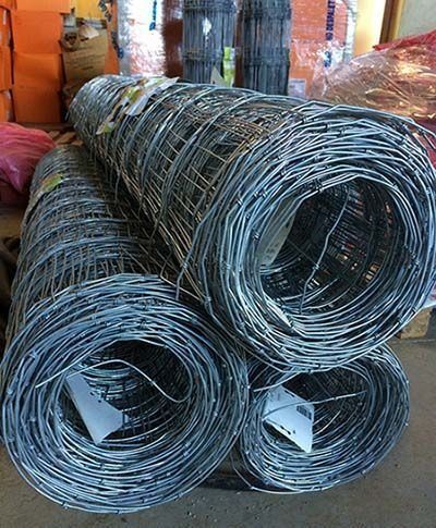 Bobinas de malla de alambre plateado apiladas en un almacén o taller, con cajas de naranjas al fondo.