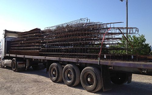 Remolque de plataforma cargado con barras de refuerzo metálicas apiladas, estacionado sobre grava bajo un cielo azul despejado.