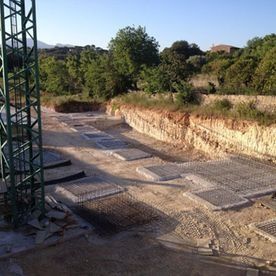 Zona de excavación con cimientos de piedra y muros de contención en construcción junto a árboles y una estructura metálica.