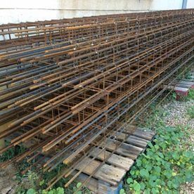 Jaulas de barras de refuerzo de metal oxidado apiladas sobre paletas de madera junto a una pared, con césped en primer plano.