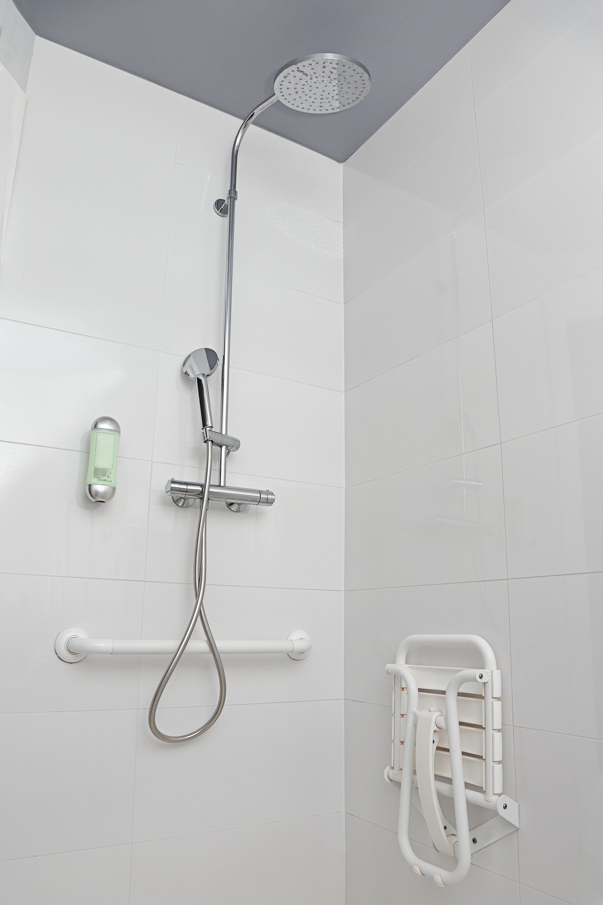 Cabine de douche carrelée blanche avec équipements d'accessibilité : barre d'appui, siège rabattable et pommeau de douche réglable.