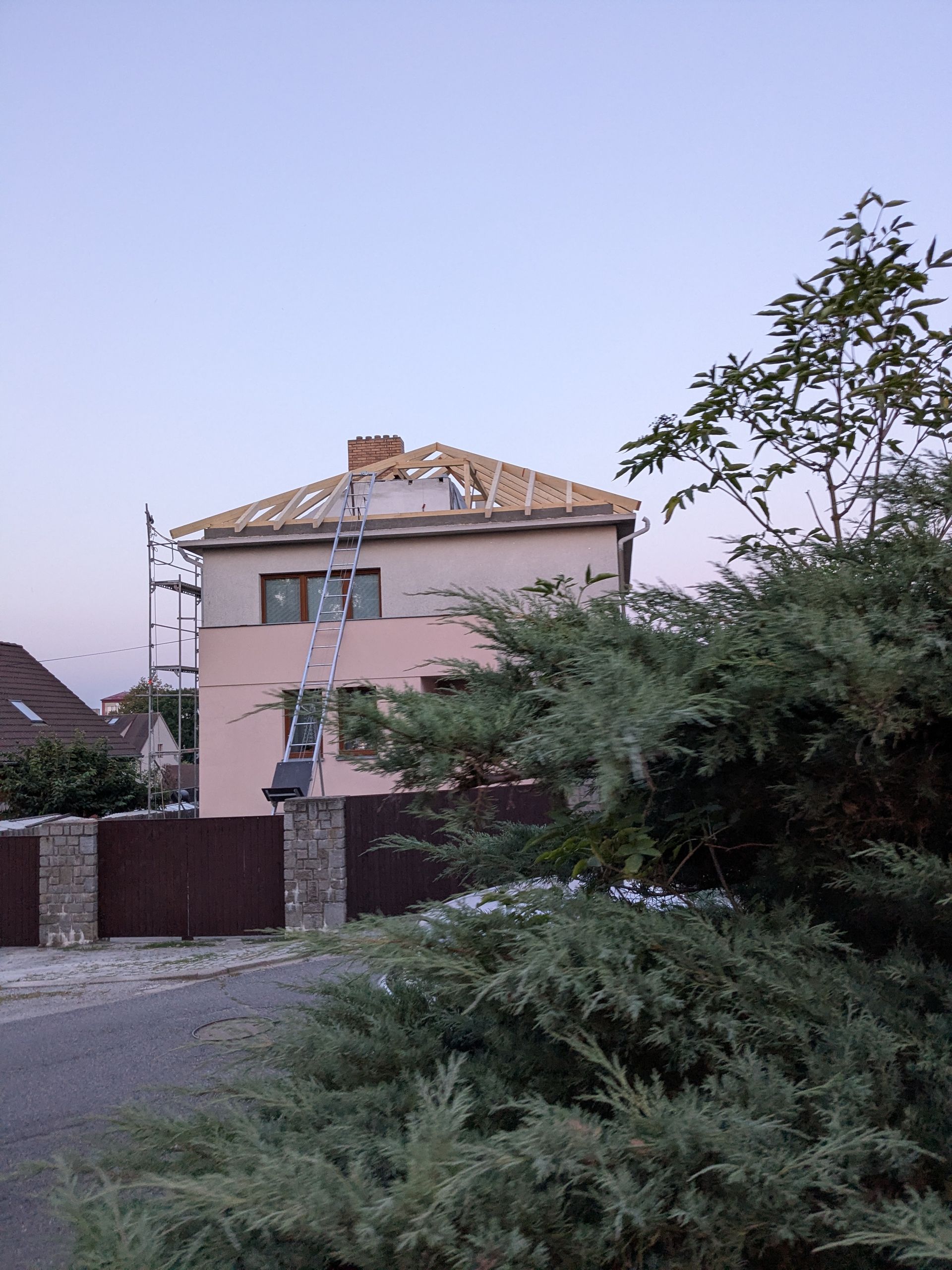 Maison rose avec échafaudages sur le toit ; clôture marron et buissons verts au premier plan, crépuscule.