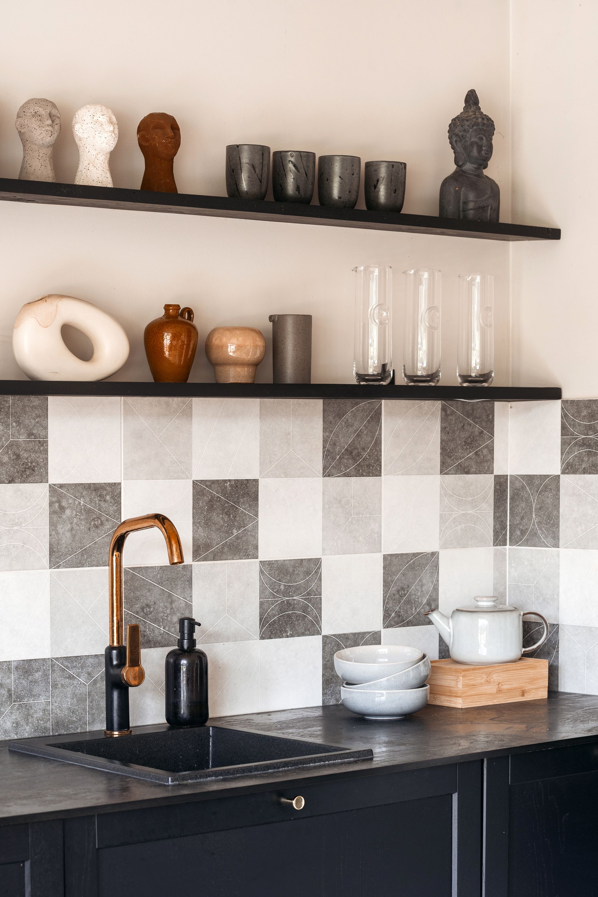 Cuisine avec plan de travail noir, robinet doré, crédence à carreaux et étagères avec objets décoratifs.