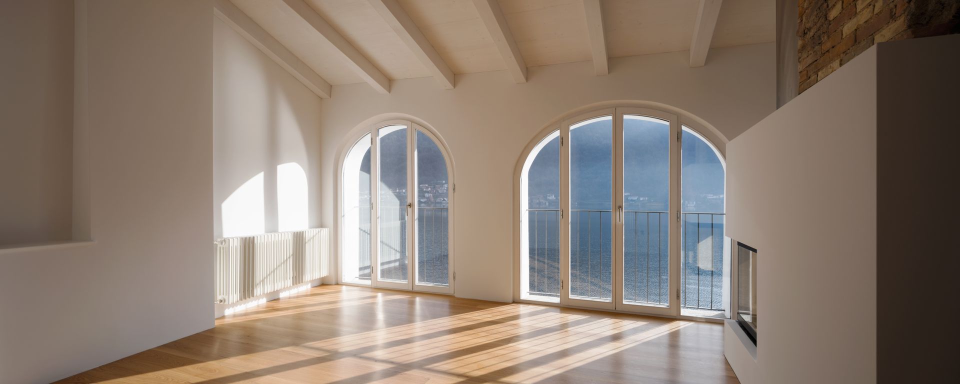 Pièce lumineuse et vide, avec des fenêtres cintrées donnant sur l'eau. Parquet, murs blancs et poutres apparentes au plafond.