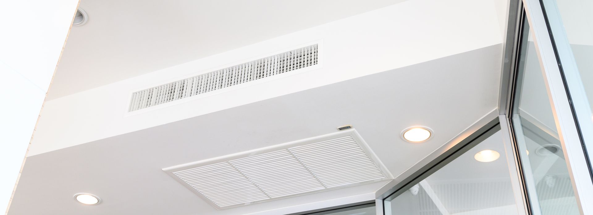 Plafond blanc avec grilles d'aération et spots encastrés. Porte vitrée sur le côté droit.