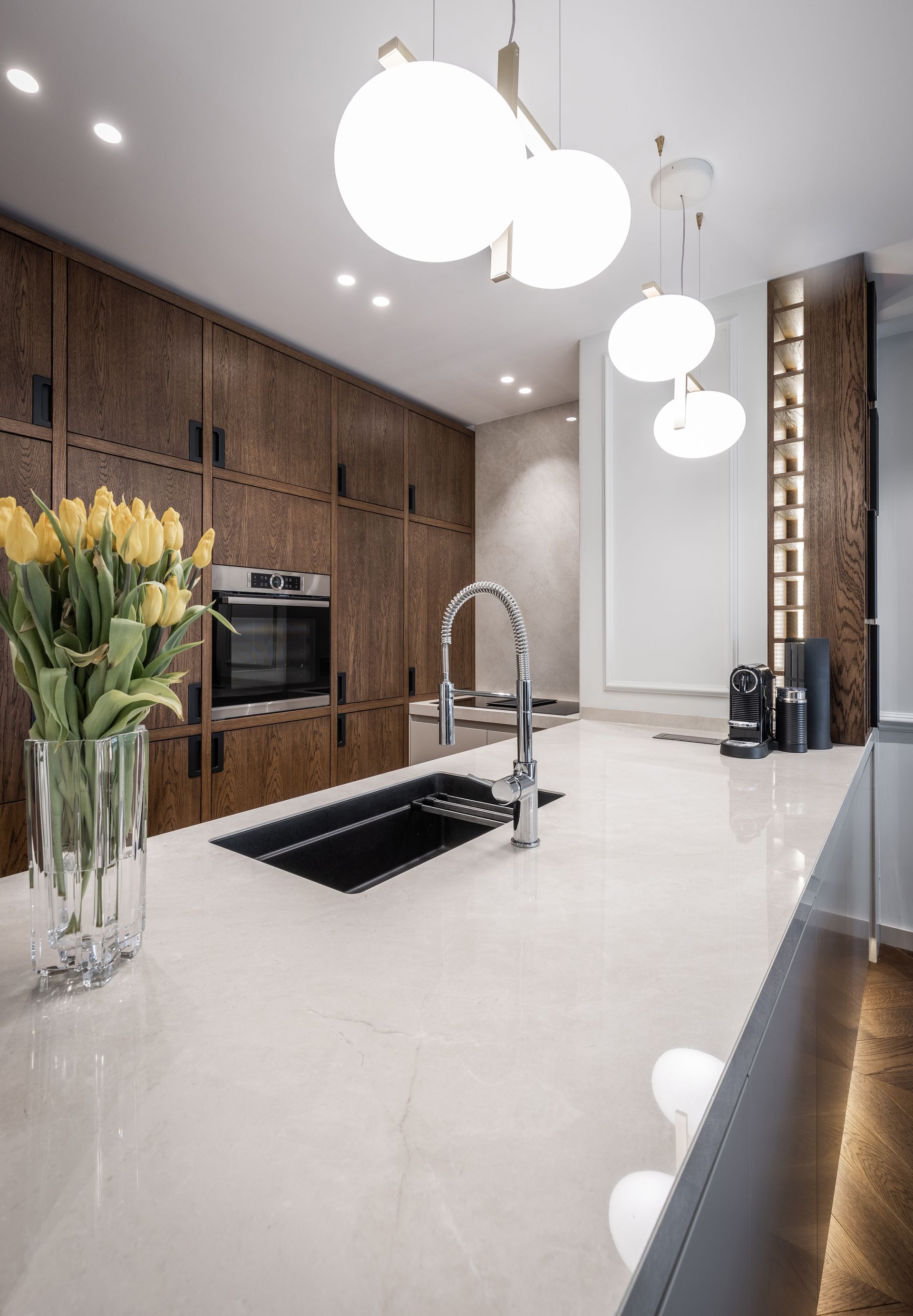 Cuisine moderne avec placards en bois, plan de travail blanc, évier noir et suspensions lumineuses en forme de globe.