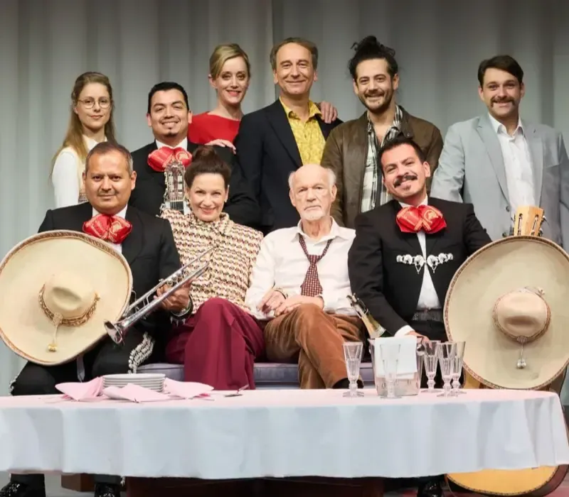 Retrato grupal de nueve personas posando disfrazadas alrededor de una mesa, con dos sentadas al frente sosteniendo sombreros.