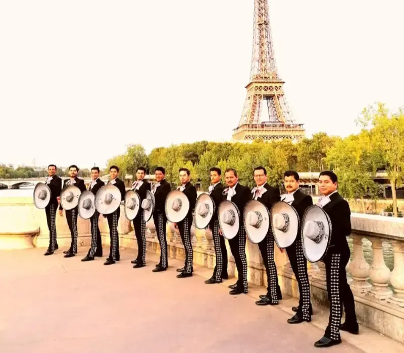 Grupo de músicos de mariachi con trajes negros y sombreros plateados, con la Torre Eiffel de fondo.