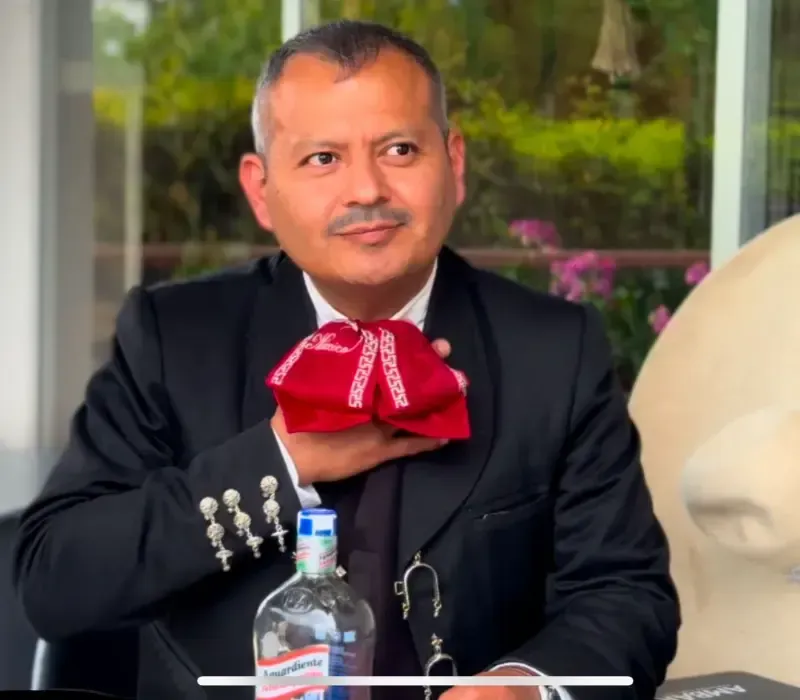 Un hombre con traje negro y pajarita roja está sentado a una mesa, mirando hacia un lado, con una botella de agua delante.