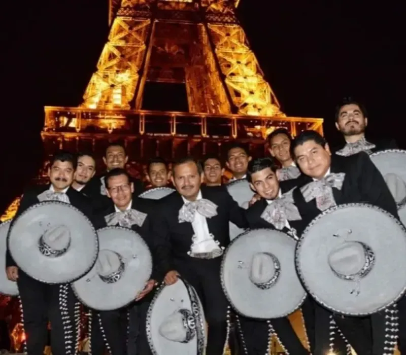 Grupo de mariachis posando frente a la Torre Eiffel iluminada por la noche, vistiendo sombreros y trajes negros.