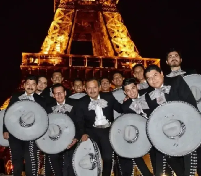Grupo de mariachis posando frente a la Torre Eiffel iluminada por la noche, vistiendo sombreros y trajes negros.