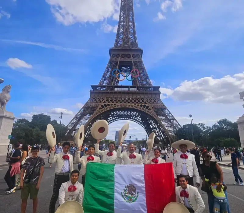 Grupo sosteniendo una bandera mexicana frente a la Torre Eiffel con artistas vestidos de blanco y sombreros cerca.