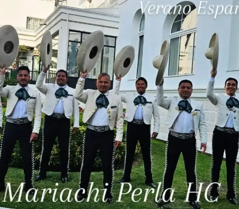 Cinco músicos de mariachi vestidos con trajes negros sostienen sombreros frente a un edificio blanco.