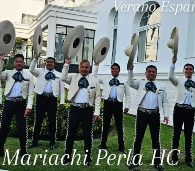 Cinco músicos de mariachi vestidos con trajes negros sostienen sombreros frente a un edificio blanco.