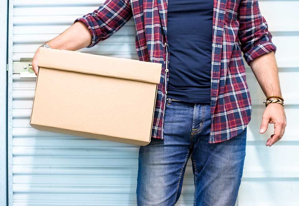 Une personne vêtue d'une chemise à carreaux et d'un jean bleu se tient devant la porte blanche d'un box de stockage, tenant un carton.