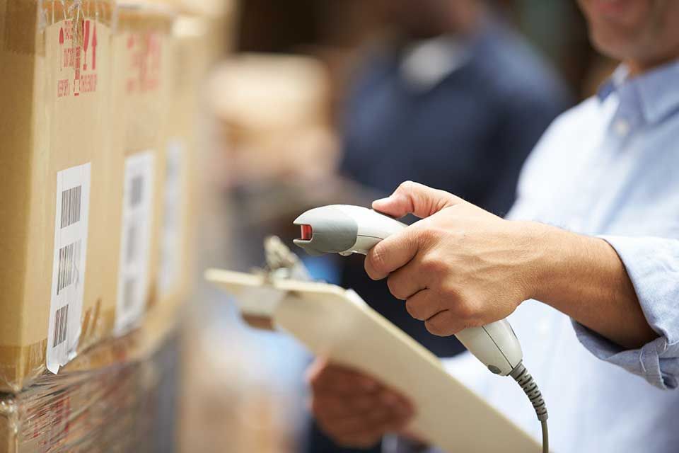 Dans un entrepôt, une personne vêtue d'une chemise bleu clair scanne un code-barres sur une boîte en carton tout en tenant un bloc-notes.