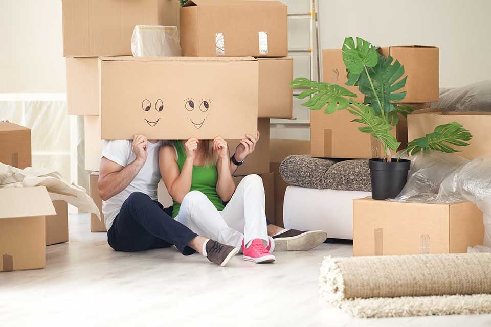 Un couple est assis par terre, entouré de cartons de déménagement, tenant une grande boîte en carton ornée d'un smiley dessiné à la main.