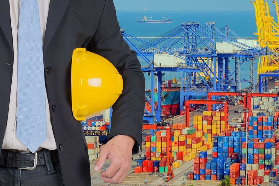 Une personne en costume et cravate tient un casque jaune devant un port de commerce animé, avec des conteneurs et des grues.
