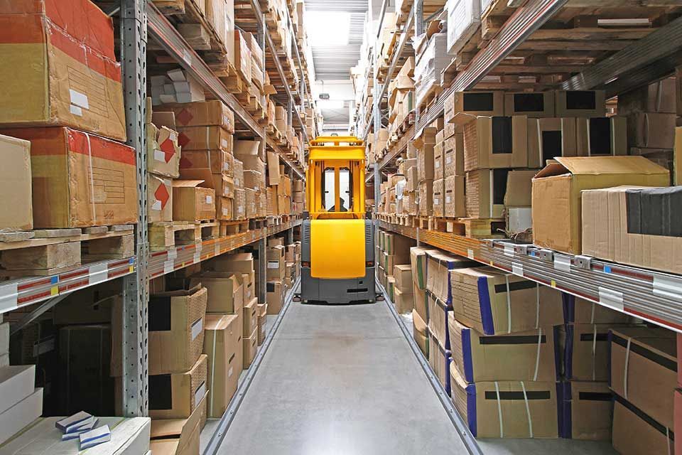 Une allée étroite d'entrepôt remplie de cartons empilés sur de hautes étagères métalliques, avec un chariot élévateur jaune au premier plan.
