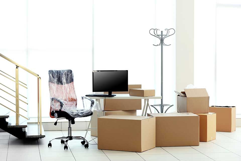 Un espace de bureau à domicile pendant un déménagement, avec une chaise de bureau emballée, un écran d'ordinateur sur un bureau et plusieurs cartons.