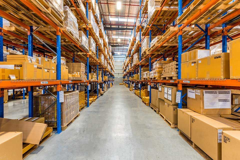 Une allée d'entrepôt bordée de hautes étagères métalliques remplies de cartons empilés sur des palettes.