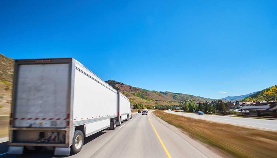 Un semi-remorque blanc roule sur une autoroute ensoleillée, longeant des collines ondulantes et des champs arides.