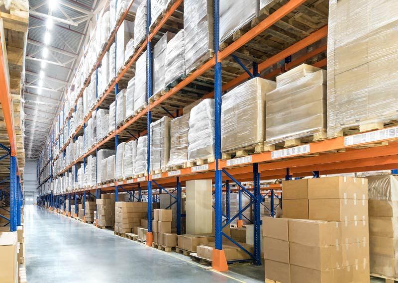 Une longue allée d'entrepôt est bordée de hauts rayonnages métalliques remplis de palettes de cartons emballés dans du plastique.