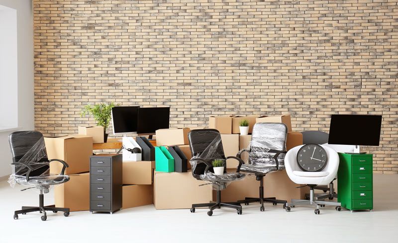Le mobilier de bureau, comprenant des chaises, des bureaux et des boîtes en carton, est disposé contre un mur de briques brun clair.