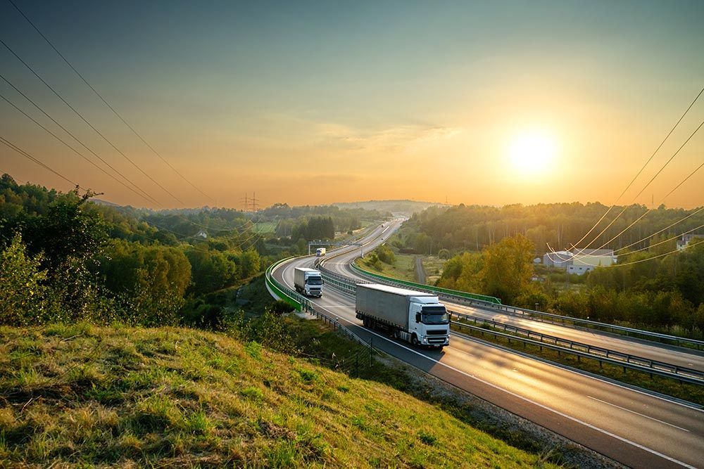 Deux semi-remorques circulent sur une autoroute sinueuse, au coucher du soleil.