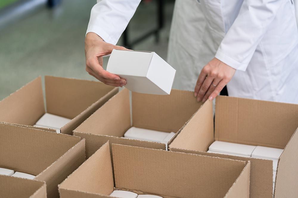 Une personne en blouse blanche place une petite boîte blanche dans un carton d'expédition.